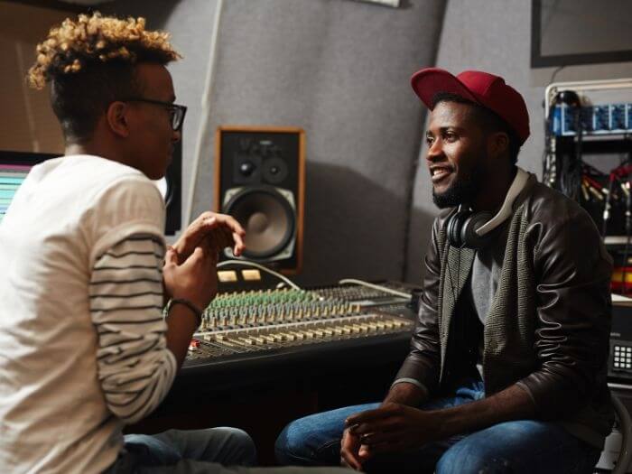 Two young Black musicians sat talking to each other in a recording studio.