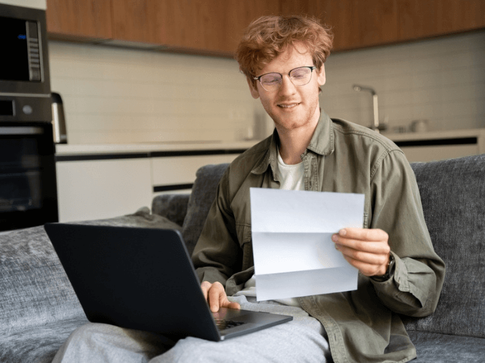 Young man sat at home on the sofa, wearing glasses, looking at a piece of paper in his hand while typing on an open laptop on his knee.