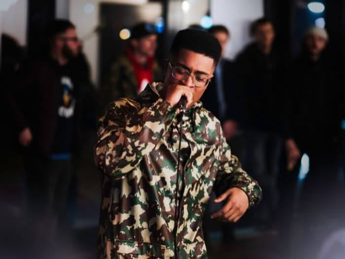 Black male rapping with microphone in camo jacket and glasses with a small crowd in the background.
