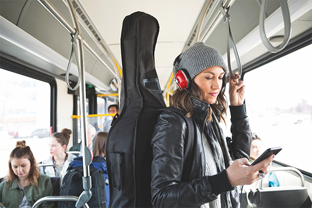 A woman standing on a bus looks at her phone. She is wearing headphones and carrying a guitar