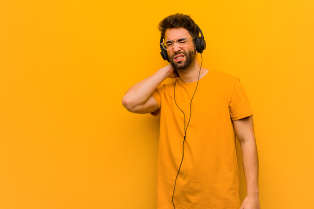 Photograph of a young man in a yellow t-shirt against a yellow background, he is wearing earphones and stretching with an expression on his face indicating he may be in pain.