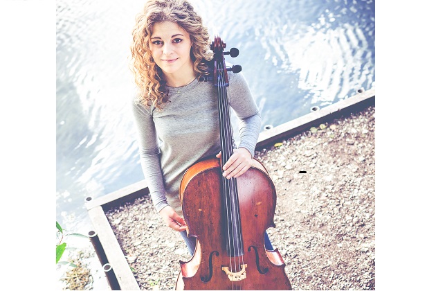 Photograph of the author, Maya Kashif. She is standing in front of a body of water, holding her cello.