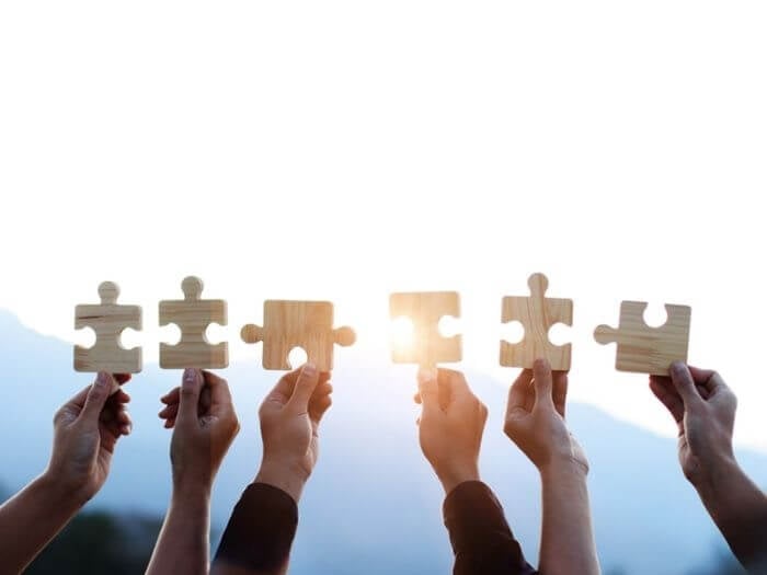 Multiple raised hands against a bright, white sky, each holding a piece of a wooden jigsaw puzzle. A teamwork, problem-solving concept.