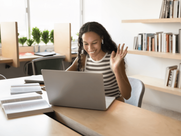 A young woman sitting at a laptop, smiling and waving to someone on her screen.