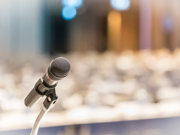 A clear shot of a microphone against a blurred background that implies there are a lot of chairs set out for audience members.