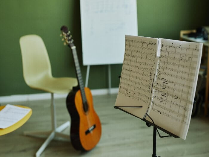 Music stand holding open music books with chairs and an acoustic guitar in the background.