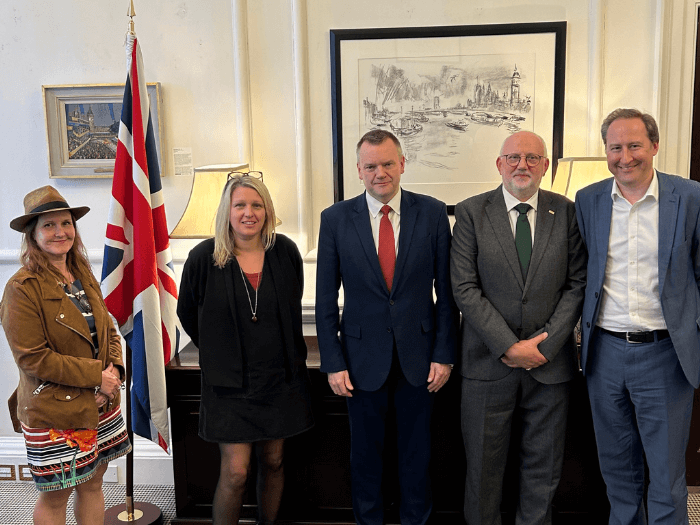 Annabella Coldrick, Naomi Pohl, Nick Thomas-Symonds, Dave Webster and Tom Kiehl smiling in front of a UK flag.