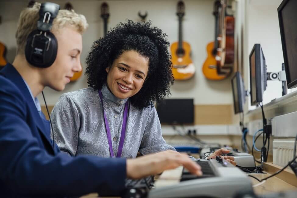 Young female teacher sat with a pupil in a music room where he plays an electronic keyboard.