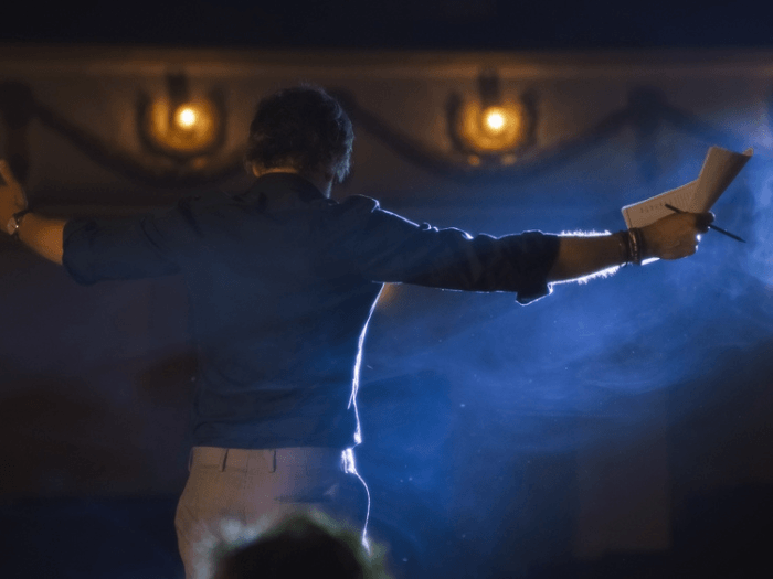View from behind a person on stage against smoke and lighting, their arms are raised as they explain something, holding a pen and paper.