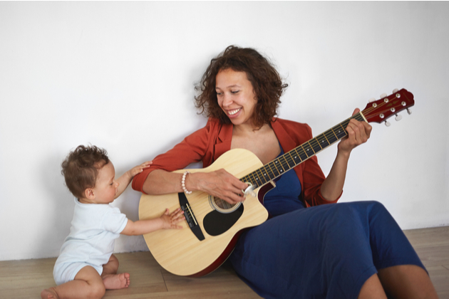 Photograph of a young mother, playing on a guitar whilst being distracted by a toddler who is reaching out to the instrument. They are both smiling.