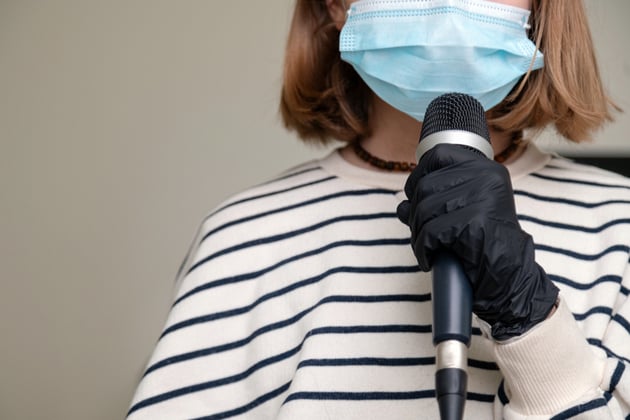 Photograph of a musician, we can't see their face but they are holding a microphone with plastic gloves on, and have a surgical mask on their face.