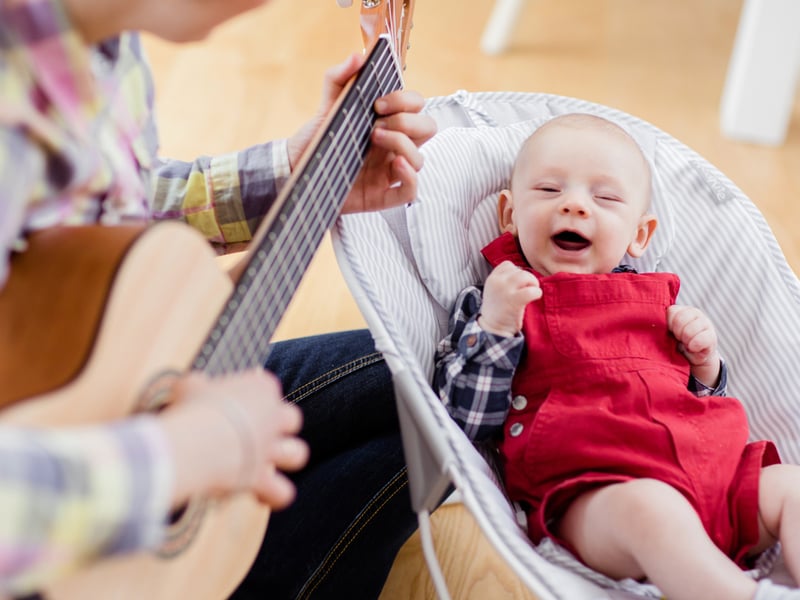 Photograph of a musician playing guitar to a laughing baby in a small baby chair.