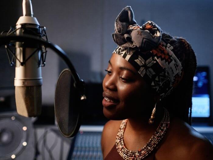 Young Black female musician singing into a studio mic with pop shield in a recording studio.