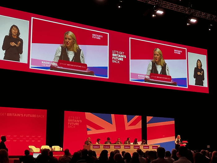 Naomi appears on the large screen at the Labour Party conference, and stands in front of a panel