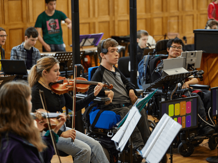 An orchestra rehearsal with young people playing various instruments, two have adaptive instruments.