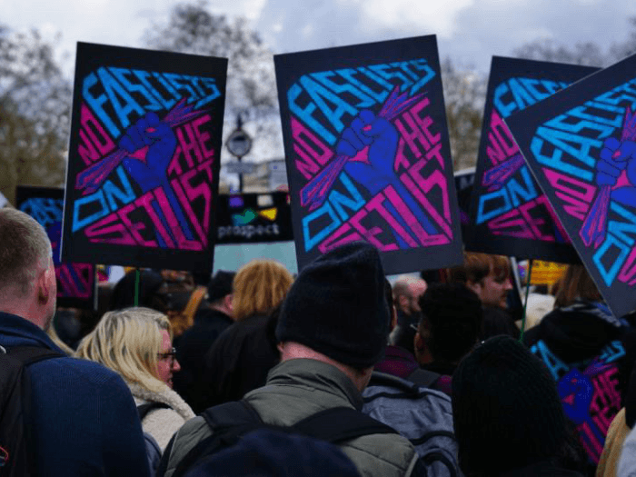 The back of a crowd of people holding black placards which show a fist holding crunched up paper with the text 