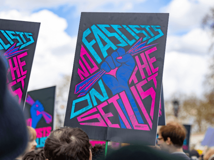 A black placard being held high in a crowd. The design is of a fist being raised powerfully in solidarity holding a piece of paper in bright blue and pink. The text says 