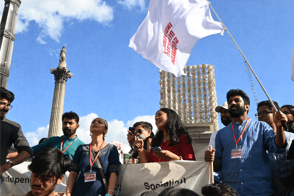 Nupur and colleagues from SFI-UK standing in a line in front of a banner on a bright sunny day with a blue sky. Some people are holding placards and Nupur is holding a microphone to address the crowd.