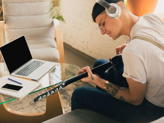 Young male musician playing guitar at home, wearing headphones in front of a laptop.