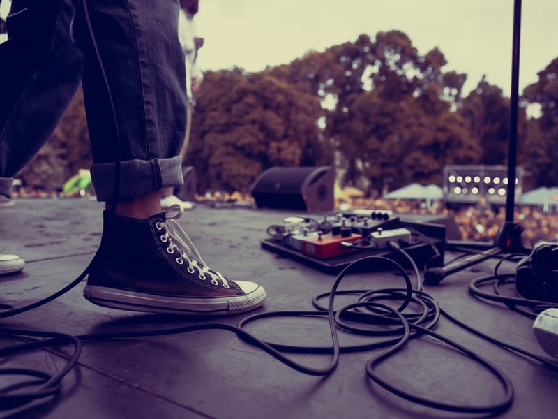 Close up of a performer at a festival, we can only see the foot of the performer in converse shoes, near a row of pedals - the rest of the photo looks out to an audience stood outdoors.