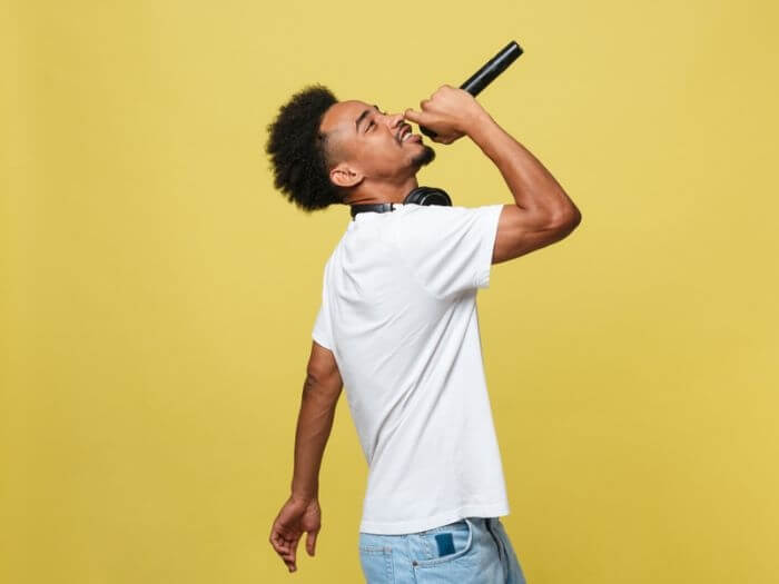 Young Black male musician, performing with microphone against a plain yellow background.