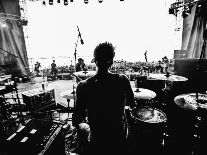 Black and white image of a band on a large stage, performing to a crowd outside. From the perspective of the drummer.