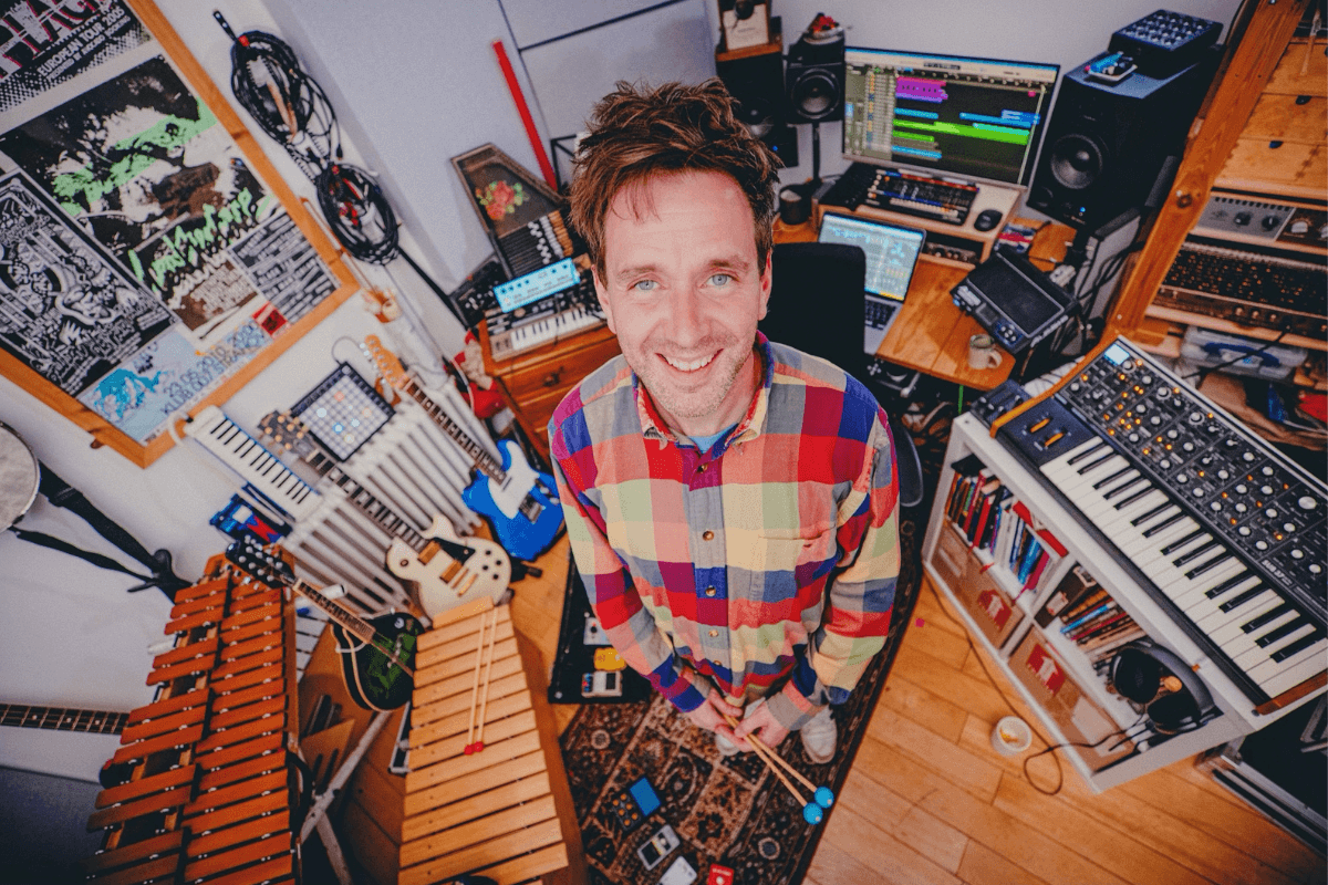 Birds eye view of Paul Russell smiling up at the camera in his home studio surrounded by equipment and instruments.