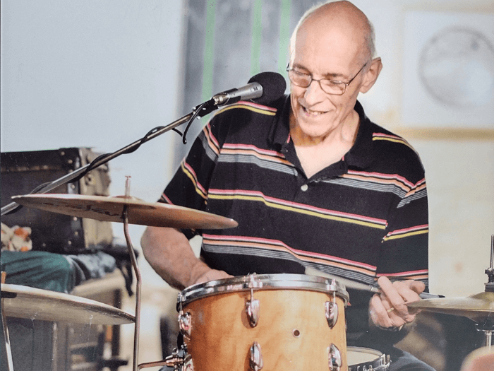 Peter smiling while playing drums and singing into a microphone.