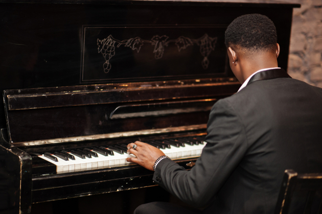 Piano player wearing a black suit seen from behind, piano player is sat at a piano.