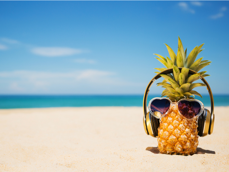 A pineapple wearing earphones and heart-shaped sunglasses is stood on a white sanded beach with blue sea and bluer sky in the background.