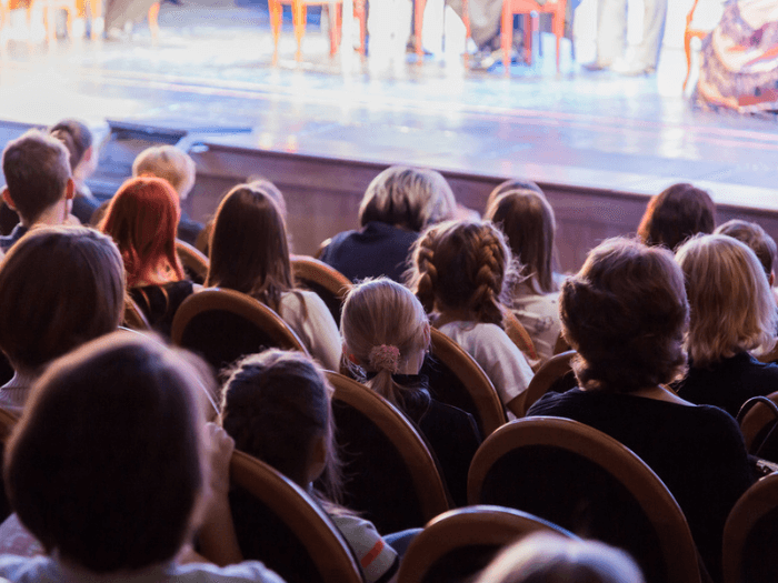 The backs of young children sat in the theatre looking at the stage.