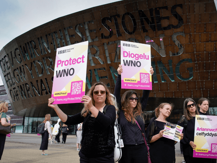 WNO orchestra players holding up placards that say 'Protect WNO'.