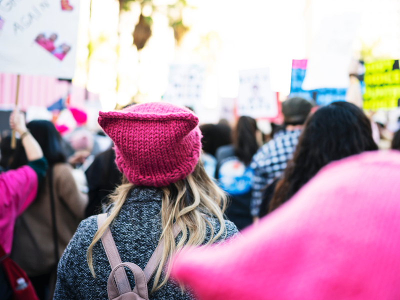 Blurred vision of a protest march seen from behind, the centre of the image is on a person with long blonde hair under a pink hat, wearing a grey coat.