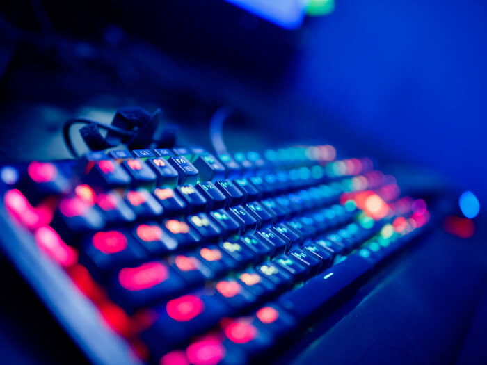A keyboard is propped up at a diagonal angle with rainbow lights highlighting the different keys, the background is dark.