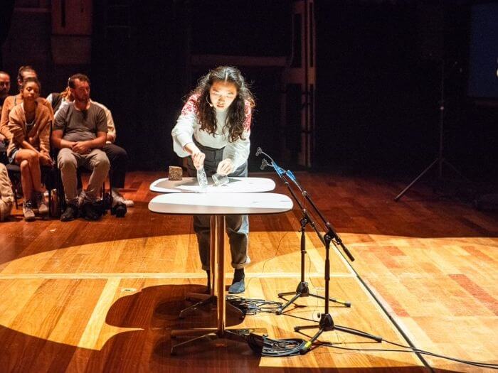 Performer Susanne Xin in front of an audience, next to a microphone and table.