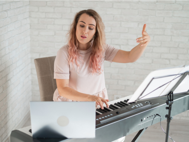Woman with pink hair sat at keyboard with left hand in the air, looking at an open laptop, teaching online music.