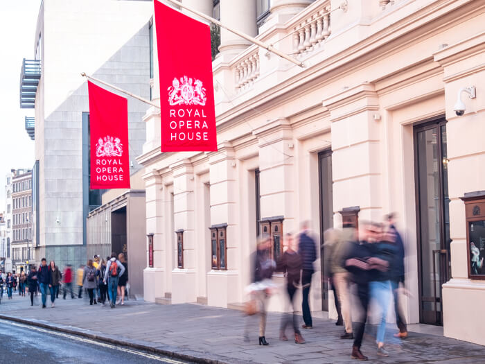 The Royal Opera House exterior showing red logo flags, with a motion blur of people outside.