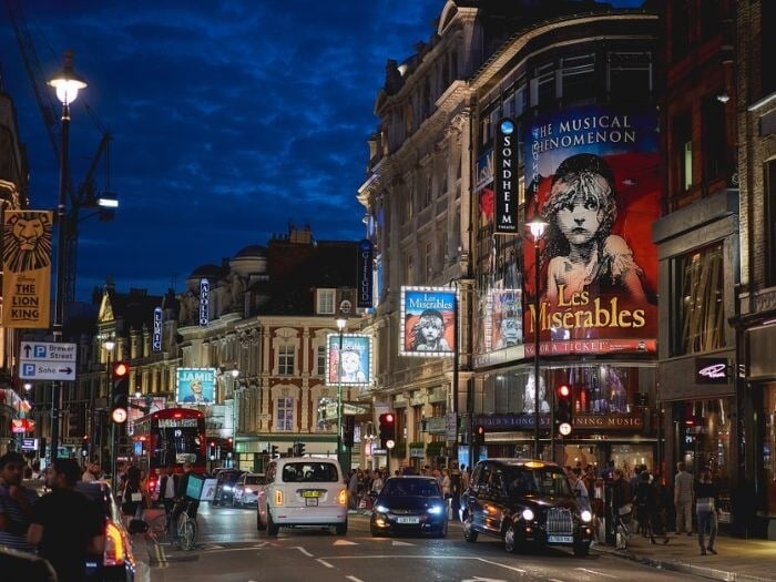 Shaftesbury Avenue London in the evening, with signs advertising different West End shows lighting up the street.