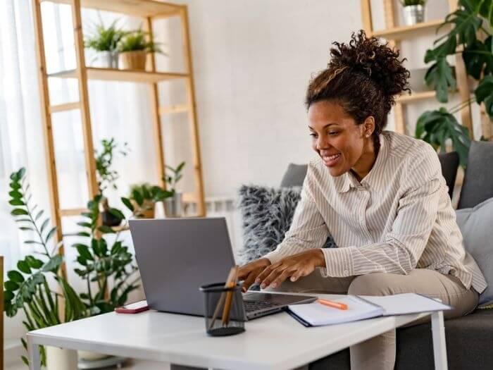 Young Black woman smiling at an open laptop at home surrounded by pens and a note pad.