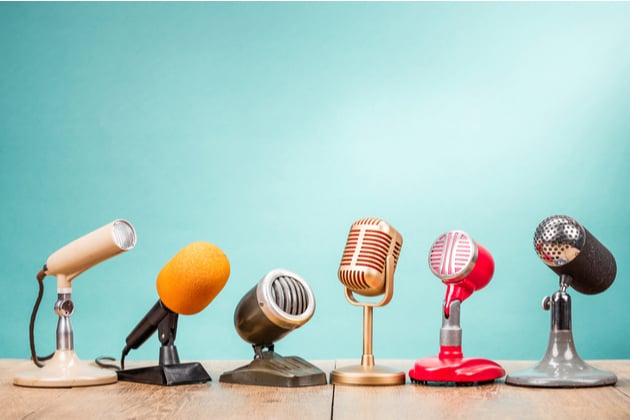 Photograph of a variety of microphones, each one a different style of vintage, set out in a row on a table. The background is a shiny blue.