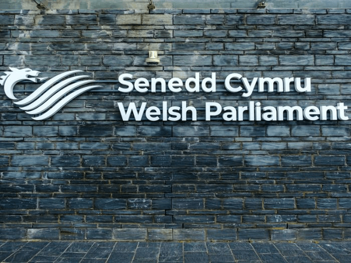 Exterior white sign on grey slate with logo saying Senedd Cymru and Welsh Parliament.