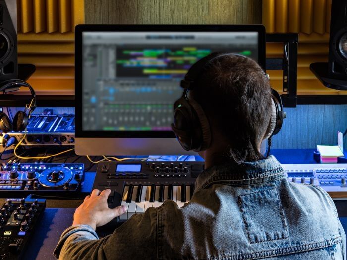 A musician sitting at a desk in a studio at a digital-audio workstation.