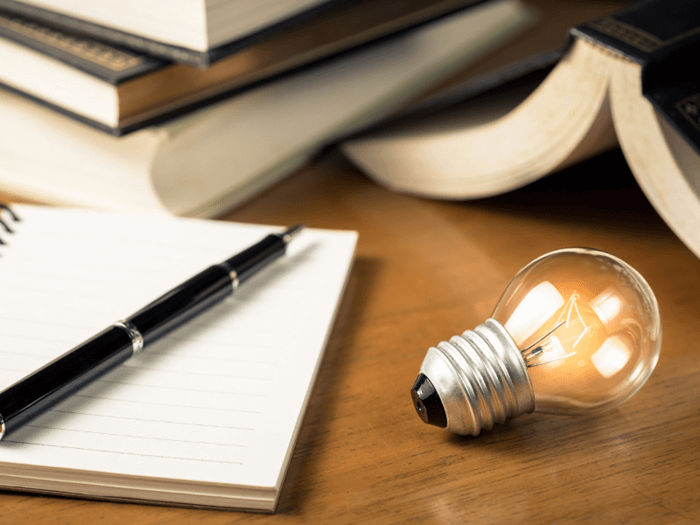 Open note pad and pen next to lit light bulb, representing storytelling and ideas
