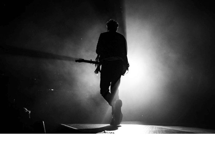 Black and white image of a man in silhouette on stage, under the spotlight with a guitar.