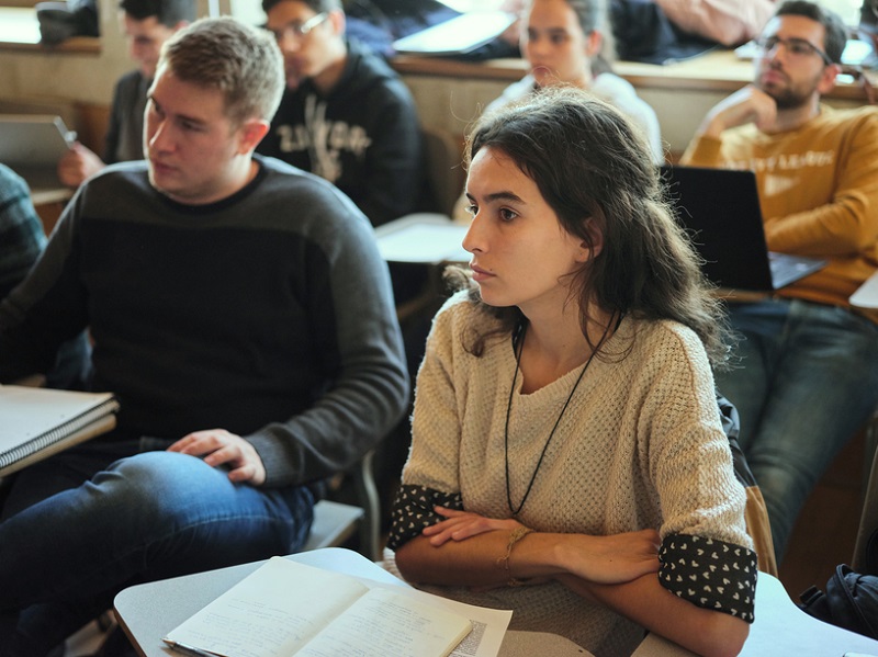 Photograph of a collection of young adults in a classroom setting, the photo focuses on one young woman facing seriously forward, with other students blured in the background.
