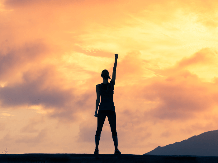 Silhouette of woman standing on a mountain/hill with her right arm in the air, the background is an orange sunrise in the clouds, representing success