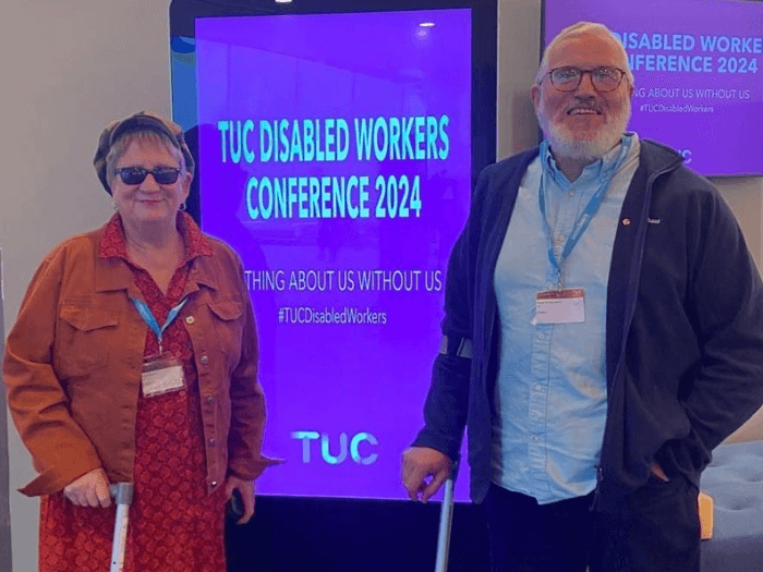 Two MU members attending the Disabled Workers Conference 2024, standing in front of a digital screen wearing lanyards.
