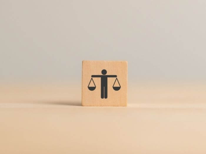 Wood cube with the figure of a person on, hands outstretched holding scales in each one which are evenly balanced.