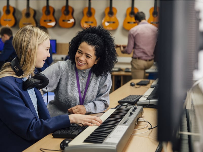 Photograph of a teacher working with a student in a classroom, the student is playing onto a keyboard and the teacher is smiling encouragingly.