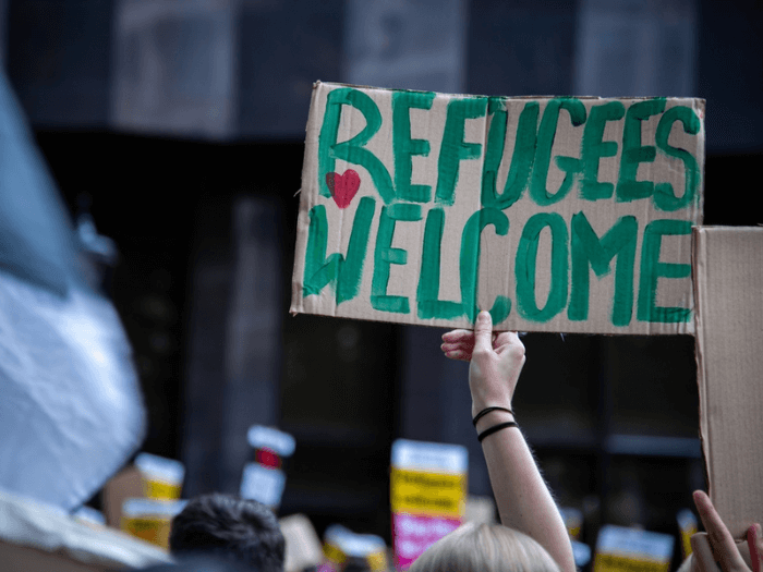 A person in a crowd holds up a handmade cardboard sign reading “Refugees Welcome” in large green letters with a small red heart. Other protest signs are visible blurred in the background.